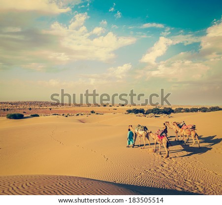 Vintage retro hipster style travel image of Rajasthan travel background - two Indian cameleers (camel drivers) with camels in dunes of Thar desert. Jaisalmer, Rajasthan, India