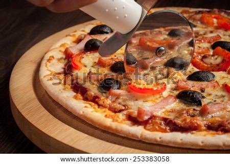 Pizza cutter (wheel) slicing ham pizza with capsicum and olives on wooden board on table