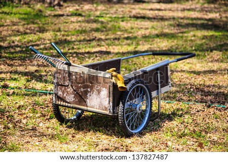A garden cart and tools with gloves hanging off the side as it sits in a lawn ready to go to work.