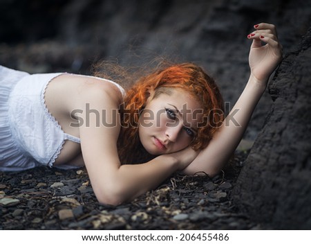 Beautiful redhead woman at the rocky beach in a white dress layed down on the gravel.
