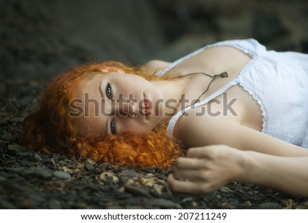 Beautiful redhead woman at the rocky beach in a white dress laid down on the gravel. Toned to warm color.