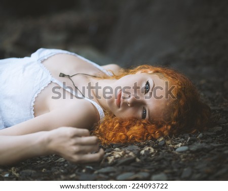 Beautiful redhead woman at the rocky beach in a white dress laid down on the gravel. Color toned image.