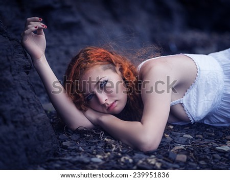 Redhead woman at the rocky beach in a white dress layed down on the gravel. Color toned image.