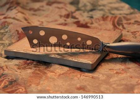 Cheese   knife on tile on brocade  table runner.  Knife has holes.