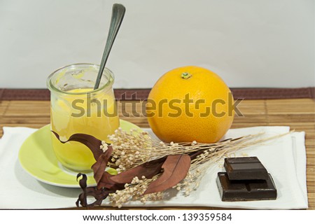 On the left side a glass of orange yoghurt with a spoon on a green plate, on the right side an orange, a few ounces of chocolate and a bouquet of dried flowers