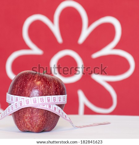 Closeup of a red apple and tape measure on a white table, in the background a red wallpaper with big white flowers