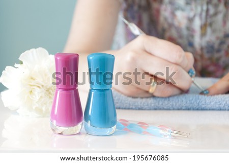 In the foreground there are two nail polishes (one pink and one blue) with manicure tools. In the background out of focus, woman\'s hands painting her nails with blue nail polish