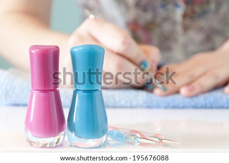 In the foreground there are two nail polishes (one pink and one blue) with manicure tools. In the background out of focus, woman's hands painting her nails with blue nail polish