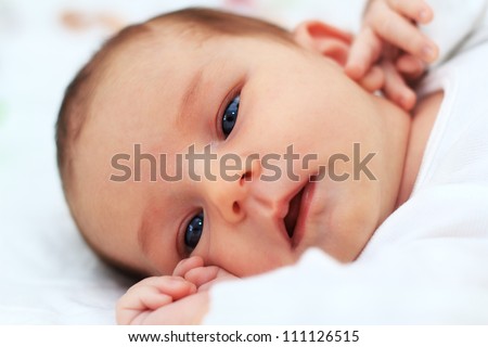 Close up of a baby girl\'s face who is holding her arms close to her face. She is dressed in white and also background is white.
