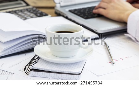 Coffee Cup at Office with Paperwork, Agenda and Businessman Working at Laptop in the Background