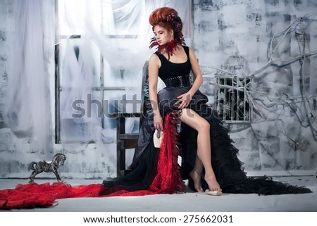 Young beautiful girl in the form of the evil fairy with red hair in a designer dress with a long skirt in unusual shoes and a necklace of feathers