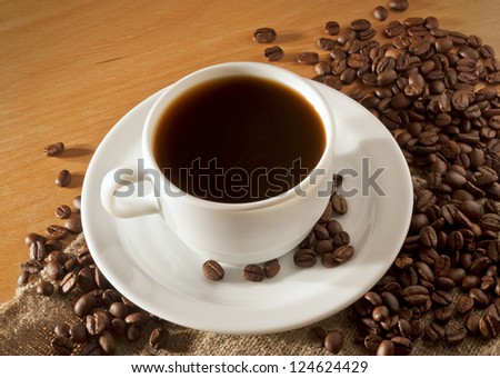 White coffee cup and saucer stands on a wooden table with a lot of coffee beans, top view