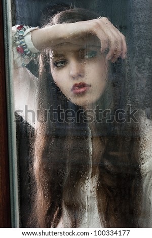 Portrait of a lovely young lady looking through glass window - Indoor in a dark cloudy day, she looks down at right and her head is resting on the right arm