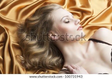 close up portrait of blond beautiful girl with well done hair style laying down on golden shining material, her face is turned in profile at left and her eyes are closed, her right hand in near the