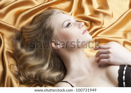close up portrait of blond beautiful girl with well done hair style laying down on golden shining material, she is turned in profile at left and looks up, her left hand is near the left shoulder