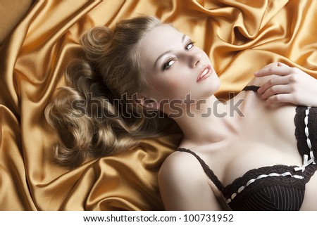 close up portrait of blond beautiful girl with well done hair style laying down on golden shining material, she looks in to the lens and smiles, her left hand is near the left shoulder