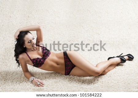 sexy brunette laying on a white carpet and wearing an elegant black lingerie, her face is turned of three quarters at left and looks in front of her, her right hand is on the head