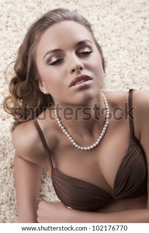 portrait of a young alluring girl in brown bra with a pearl necklace laying on a white modern carpet
