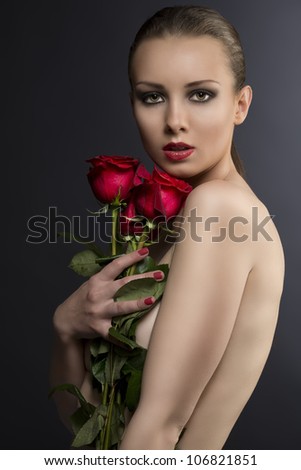 pretty blonde girl\'s low key portrait with three red roses, her body is turned in profile,  looks in to the lens and she takes the roses near the face