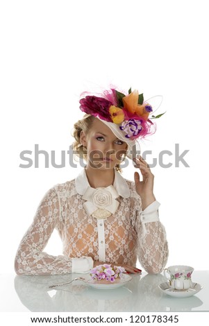 blonde girl with floral hat and vintage shirt behind the table with cake and cup of tea, she is in front of the camera and looks in to the lens