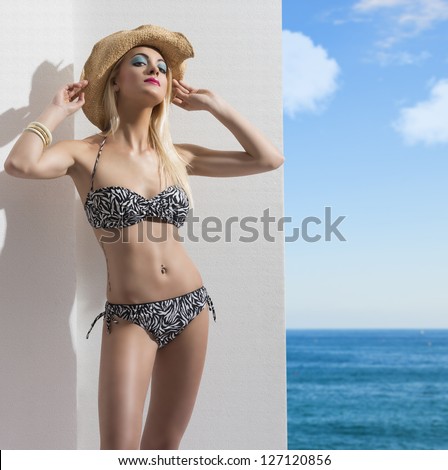 sexy blonde girl in bikini with summer hat on the head and tattoo on the hip. Blue sky and sea in background