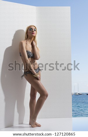 beautiful girl in summer time wearing bikini, posing near white wall with tattoo on the hip. Sea, blue sky and some boats on background