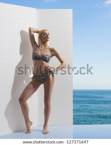 cute blonde lady in summer time with sexy bikini in fashion pose near white wall. Blue sea and clear sky on background