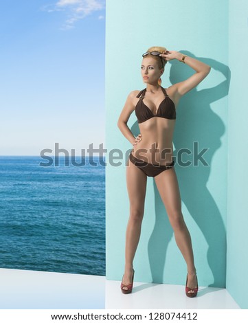 very pretty blonde girl posing near blue wall wearing cute nikini, heels and sunglasses. Sea and sky on the background