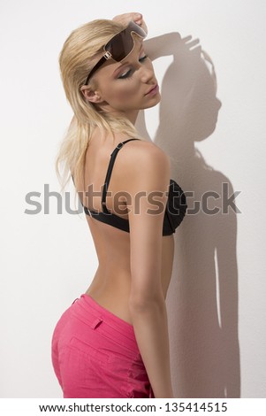 glamour summer portrait of sexy blonde girl with wet blonde hair, bra, shorts and sunglasses. She showing her back near white wall