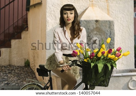 sensual brunette pin-up girl sitting on bicycle with some colorful tulips in the basket and wearing short skirt and pink shirt