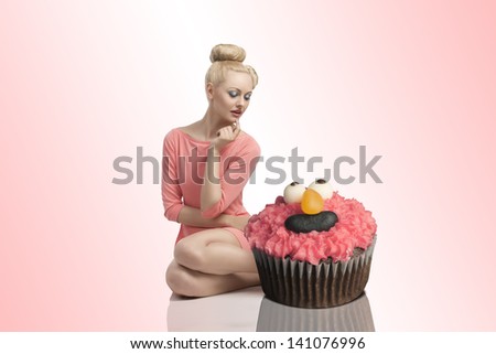 pretty blonde woman sitting near two big cupcakes with colorful make-up and creative hair-style. Wearing pink dress