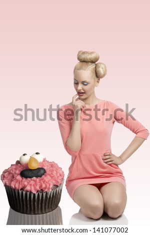 pretty blode girl with colorful make-up and creative hair-style kneeling on the floor with big cupcake . Wearing pink dress