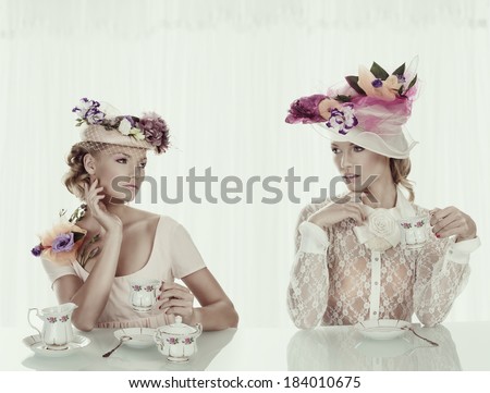two blond girl friends taking a cup of tea with old fashion flower hat in white background