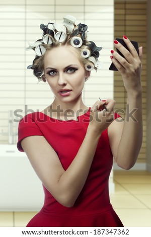 very pretty female posing in funny shoot with hair rollers and red dress. She make-up herself and putting lipstick with brush