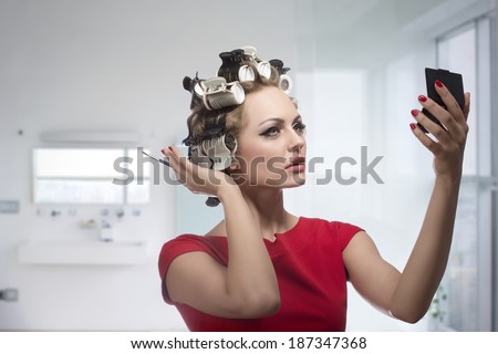 Blonde lady taking care of her style and checking that in the little mirror. Applying make-up with brush and styling her hair with curlers, wearing fashion red dress