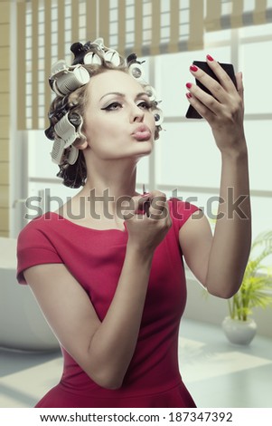sensual girl applying make-up and looking in the mirror in her hand. Styling her hair with curlers and wearing red dress. Preparing herself for party