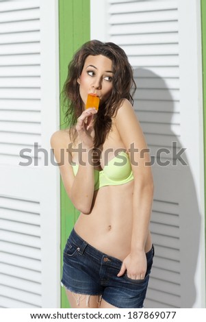 sexy young brunette girl with curly hair posing in colorful bikini with jeans shorts and eating ice lolly