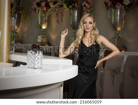 pretty woman in black evening dress , with hair style posing near a restaurant bar , looking in camera .