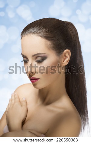 Portrait of beautiful, young woan with long, straight, brown hair clipped in ponytail, she has strong make up and she is posing on grey background, she is looking down.