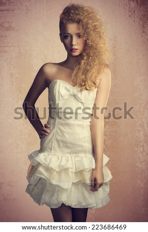Young, blonde, pretty girl with curly hairstyle and romantic look. She is wearing white, gorgeous dress with a loop and colorful makeup. She is looking at camera.