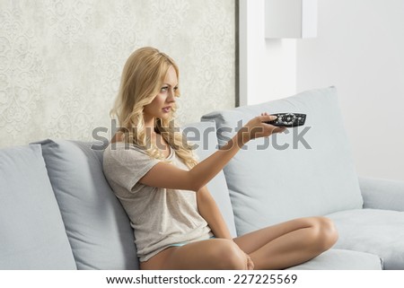 charming blonde female sitting on comfortable sofa with nude legs and panties, watching television and changing channel with remote control