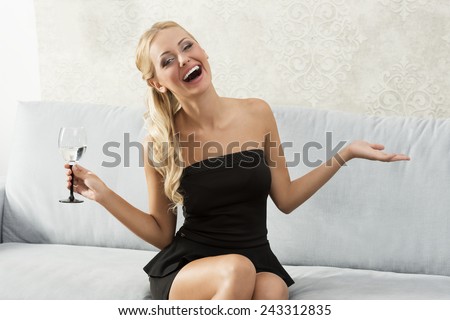 Beautiful, blonde woman is sitting on the sofa, and laughing with a glass in her hand. She wears black dress.