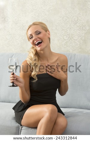 Elegant, attractive, blonde woman in black dress is sitting on the sofa and holding a glass. She is laughing.