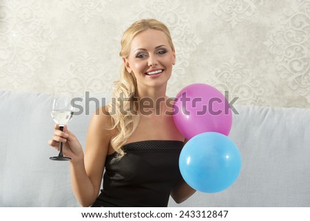 Smiling, pretty, blonde woman on party is sitting on the sofa, holding glass, and balloons. She wears black dress.