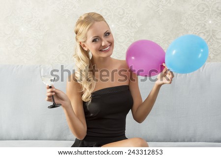 Beautiful, happy, blonde woman on party is sitting on the sofa, holding glass, and balloons. She wears black dress.