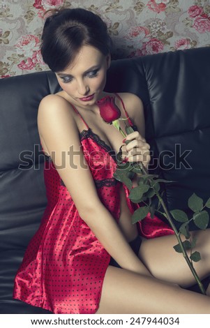 Beautiful, sexy, pretty valentine woman with brown hairstyle is sitting on the sofa in nice outfit and holding a rose.