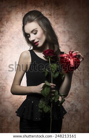 Young, beautiful, romantic, elegant girl in black dress is holding valentines present like rose and heart. Her makeup is perfect.