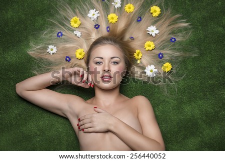 creative spring portrait of sexy woman lying on green grass with colorful flowers in the long smooth hair, covering her naked breast and looking in camera