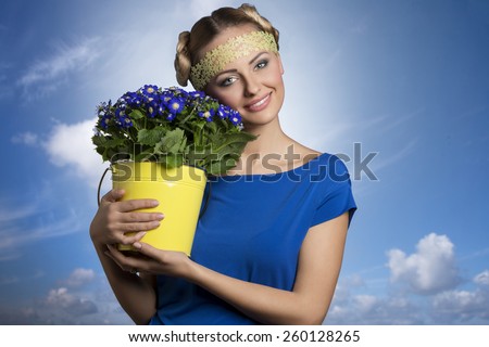 Pretty, fresh, natural, blonde woman in blue t-shirt. She is holding bucket of blue flowers. She has got funny hairstyle.