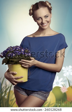 Pretty, natural, blonde, young woman in blue T-shirt is holding bucket of blue flowers. She has got nice hairstyle and make up.